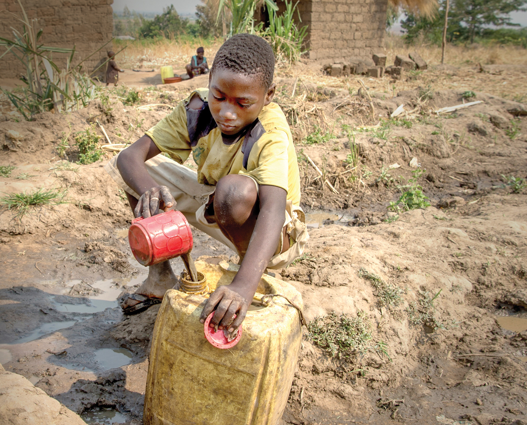A child in a yellow shirt and shorts fills a yellow jerrycan with dirty water from a small red container.